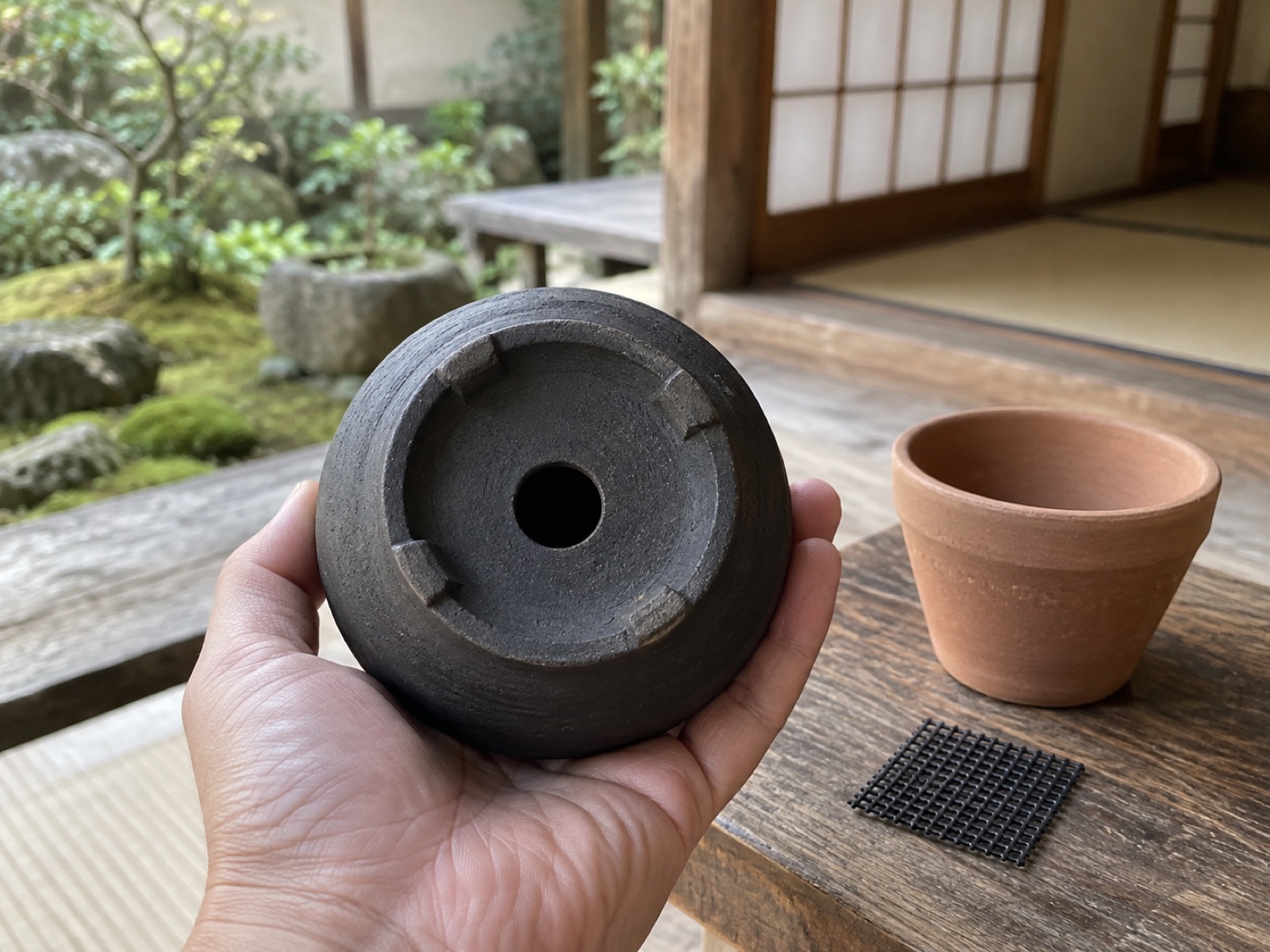 黒い焼き締めの鉢を裏返して手に持ち、鉢底の大きな排水穴を見せている手元。横に素焼き鉢と鉢底ネット